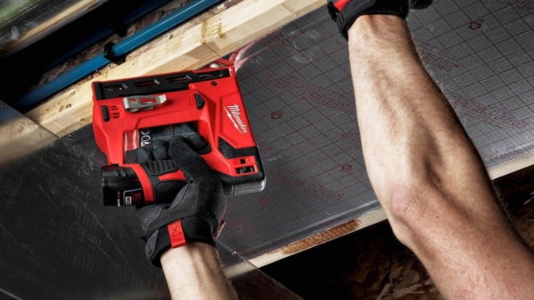 A worker wearing safety gloves using a Milwaukee crown stapler on ceiling joists