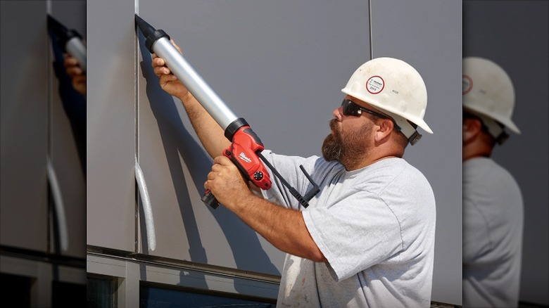 A worker wearing a hard hat using a Milwaukee adhesive gun on a gap in an exterior wall