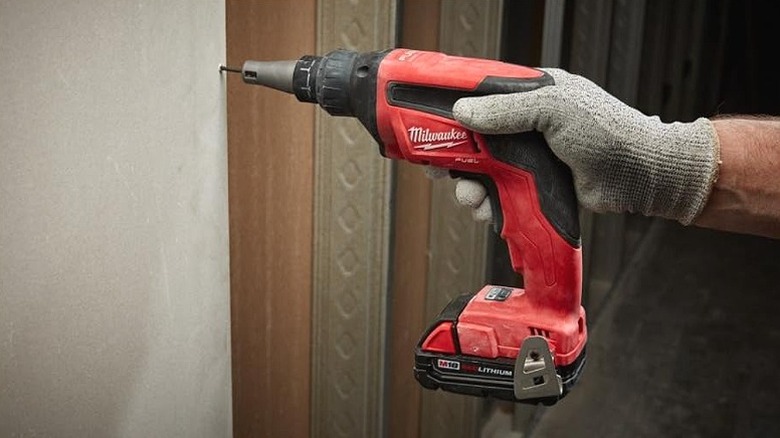 A worker wearing safety gloves using a Milwaukee drywall screw gun on a wall