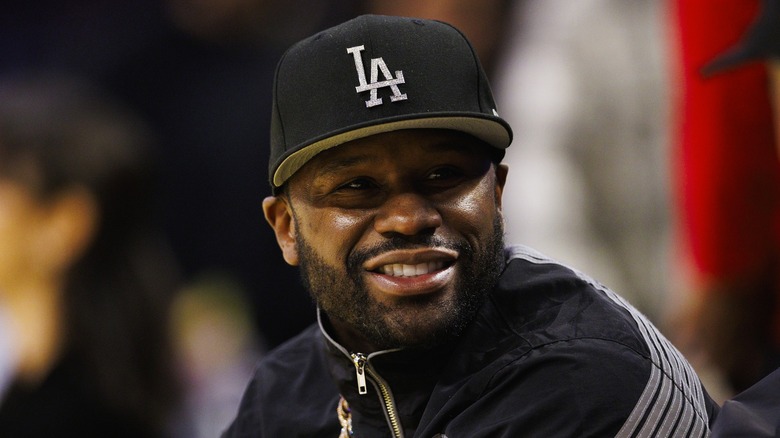 Floyd smiling at someone while wearing a black jacket with white stripes and a cap with "LA" on it.
