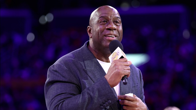 Magic Johnson speaking while holding a Lakers microphone, dressed in a grey jacket and white shirt.