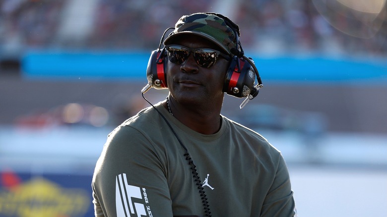 Michael Jordan wearing a grey Jordan jacket, a camo cap, black shades, and a communication headset.