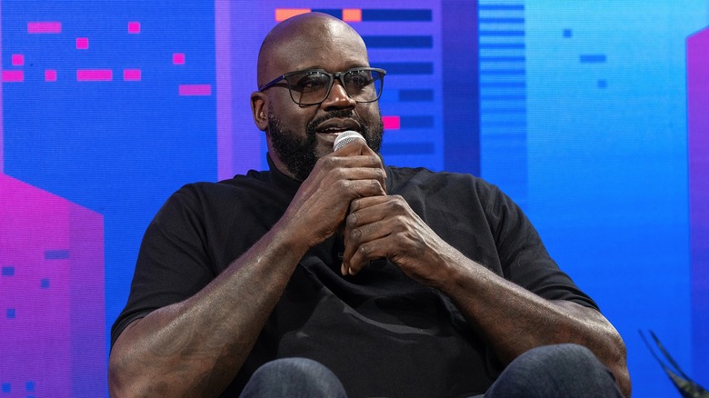 Shaq seated at a conference, speaking into a microphone, dressed in a black shirt, jeans, and glasses.