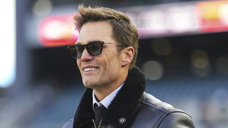 Tom Brady looking sharp in a black jacket and sunglasses, smiling confidently.