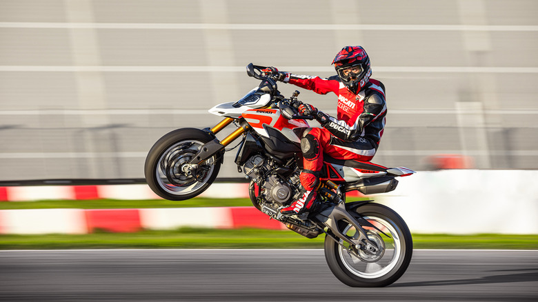 A rider in full Ducati-themed gear and helmet popping a wheelie on the new Ducati Hypermotard V2 on a track.