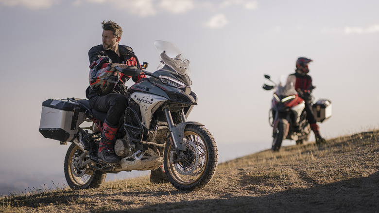 A rider with his helmet off sitting on a stationary Ducati Multistrada V4 rally, while another is out of focus in the back.