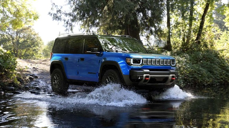 A blue 2026 Jeep Recon Trail-Rated EV SUV moving into a small stream while splashing water about in a forest.