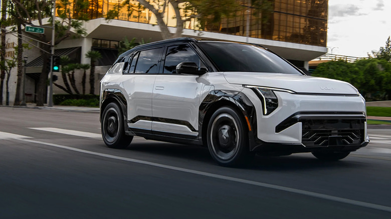 A silver 2027 Kia EV3 moving from left to right down a road with a modern glass-fronted office building behind it.