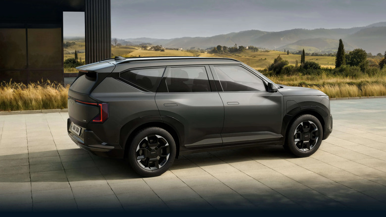 A slate gray Kia EV5 for the 2025 model year, shown from the back-right profile, parked on concrete with a field in the backdrop.