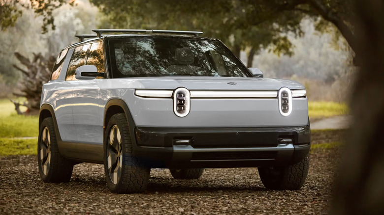 A gray Rivian R2 electric SUV standing off-road in the shade of several trees, among fallen leaves.