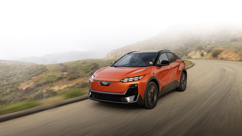 An orange 2026 Subaru Uncharted EV SUV moving at speed down a road on a hill in the day.