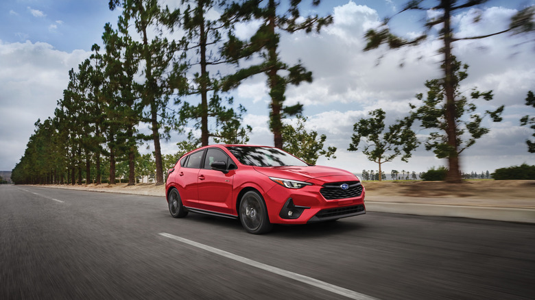 A red 2025 Subaru Impreza driving down a tree-lined road on a cloudy day.