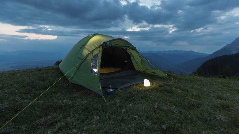 A tent is pitched on a grassy hillside with a lantern illuminating the entrance area.