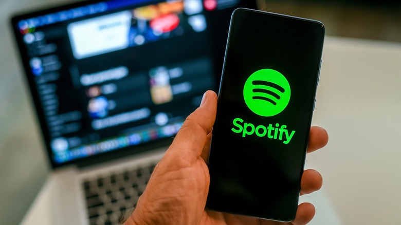 Close up view of hand with smartphone and Spotify ​logo on display. Laptop on background.
