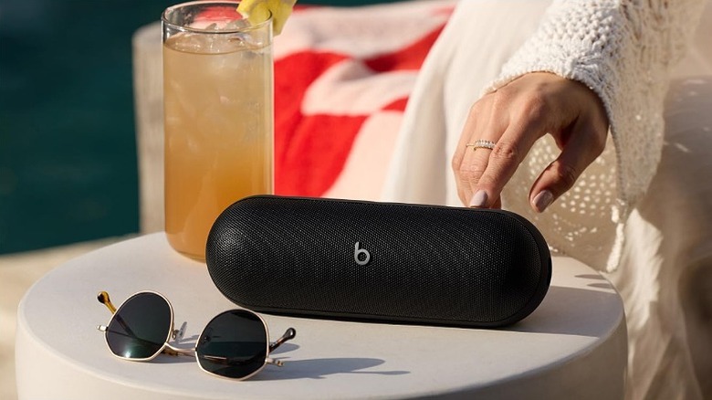 Woman pressing a button on a black Beats Pill placed alongside a glass of juice and sunglasses.