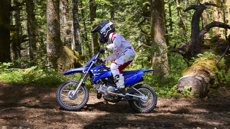 An action shot of a young rider on a Yamaha TT-R110E dirt bike in a forested off-road area.