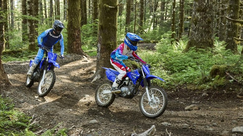 Two young riders in protective gear and blue helmets moving down an offroad trail on blue Yamaha TT-R125LE bikes.