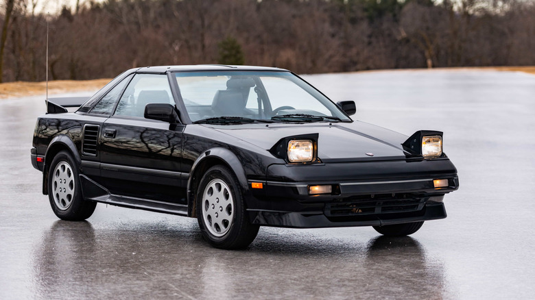 Black 1989 Toyota MR2 with headlights up parked outdoors on stone plaza with trees in background