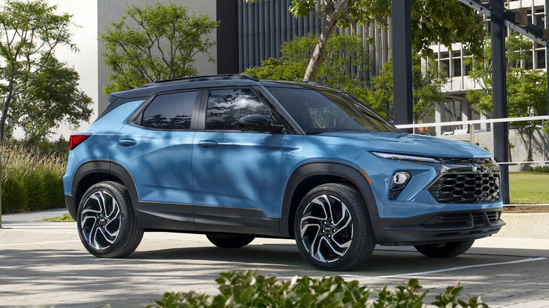 A blue 2026 Chevrolet trailblazer parked in front of modern office buildings with some trees in the background.