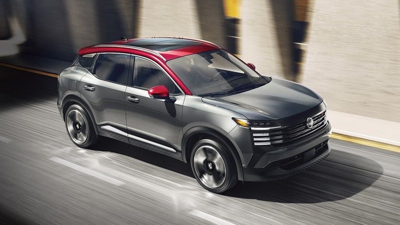 A 2026 Nissan Kicks in dark metallic gray with a two-tone red roof moving at speed down an asphalt road.