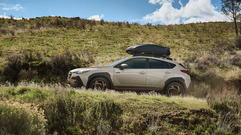 A light-colored 2026 Subaru Crosstrek going off-road while equipped with a black roof box, grass all around it.