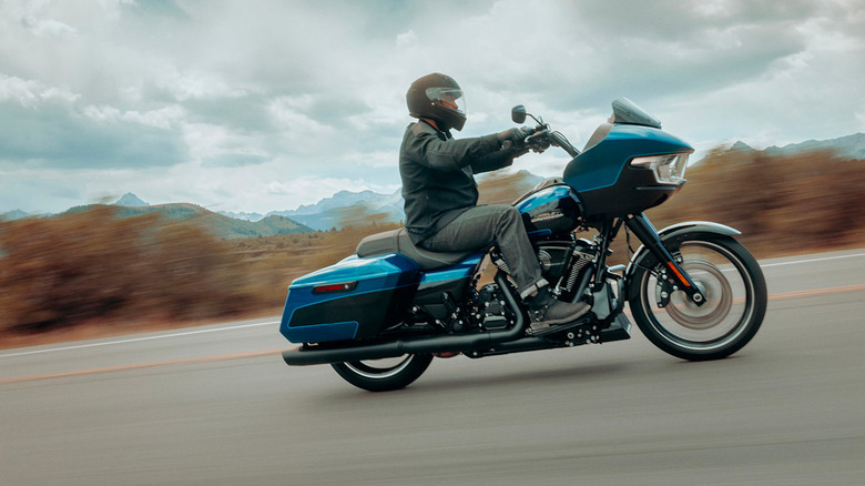 A rider in a leather jacket and black helmet riding a blue 2026 Harley-Davidson Road Glide down a highway.
