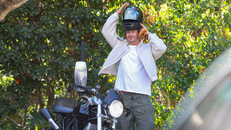 Orlando Bloom putting on a helmet next to a motorcycle