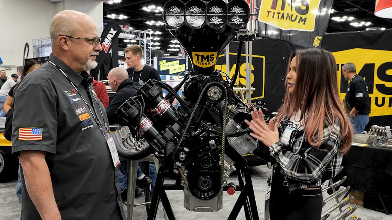 Two people standing next to a HEMI NHRA Top Fuel dragster engine mounted on a rack.