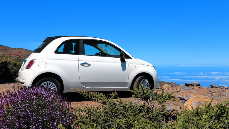 A cream Fiat 500 parked on the side of a coastal mountain, clouds in the background.