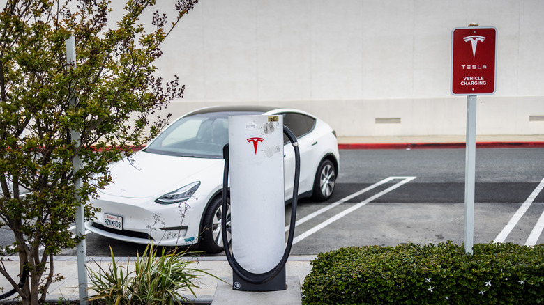 A white Tesla Model S is seen parked at a supercharger with a white building in the back.