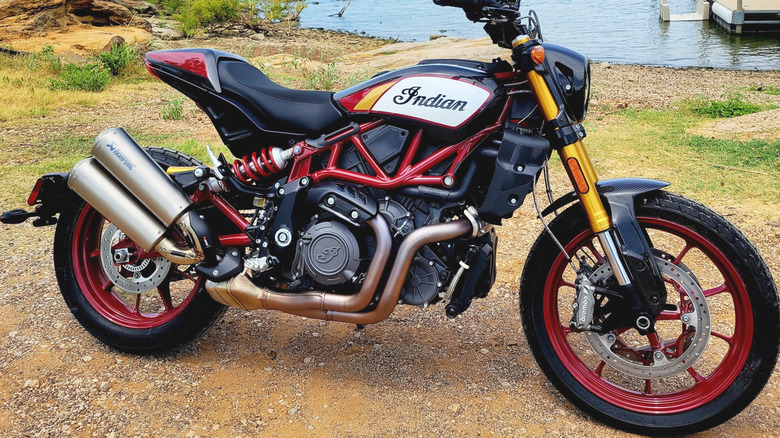 A red and black Indian FTR 1200 naked motorbike parked on sand near a beach, with the water visible in the back.