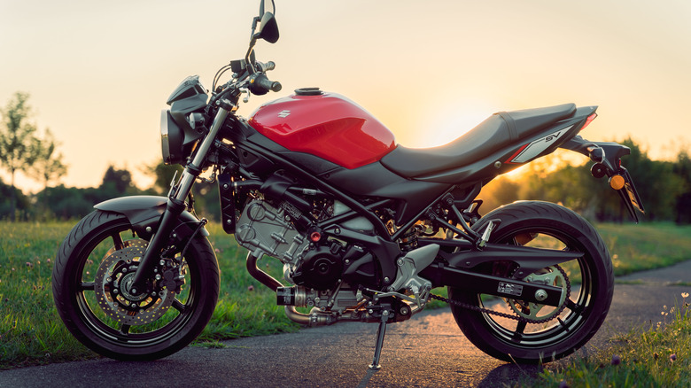 A black and red Suzuki SV650 parked on a road with grass in the background with the setting sun visible in the frame.