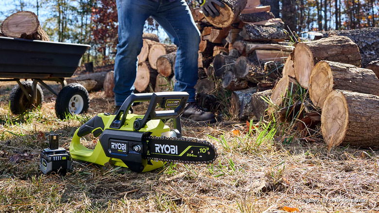 A Ryobi chainsaw sitting on the ground next to a man stacking logs.