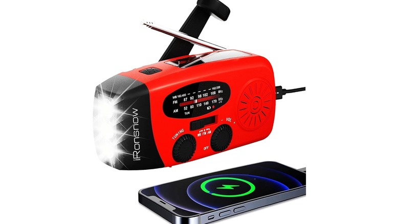 A red and black radio with antenna and flashlight, next to a charging smartphone, against a white background