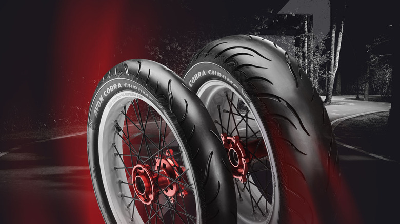 Two Avon Cobra Chrome motorcycle tires with red hubs and spoke wheels displayed against a dark, curving road backdrop with a red glow highlighting the tires.