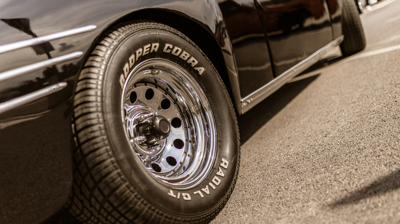 Close-up of a Cooper Cobra Radial G/T tire mounted on a shiny chrome wheel, attached to a classic black car parked on asphalt.