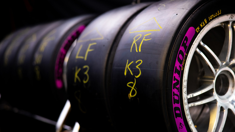 Stack of Dunlop racing tires with yellow writing, showing a close-up of one tire mounted on a silver race wheel.