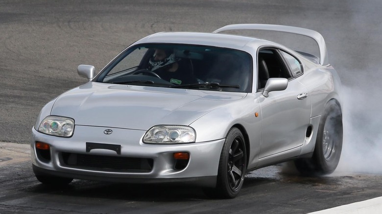 Silver Toyota Supra MK4 performing a burnout on a drag strip, with smoke rising from the rear tires and a driver visible inside.