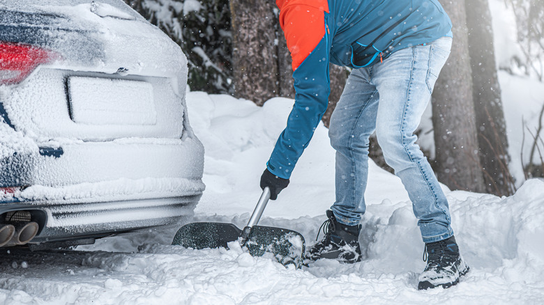 A man clearing snow from the back of a car stuck in a snowdrift using a shovel.