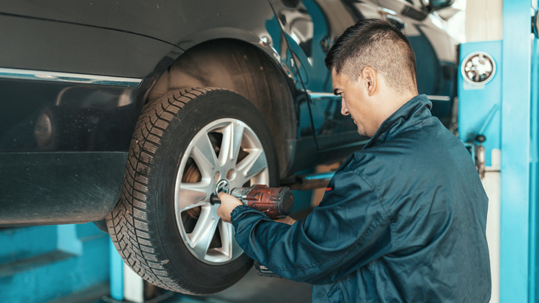A young mechanic in uniform crouching next to a tire on a car that is lifted up on a jack.