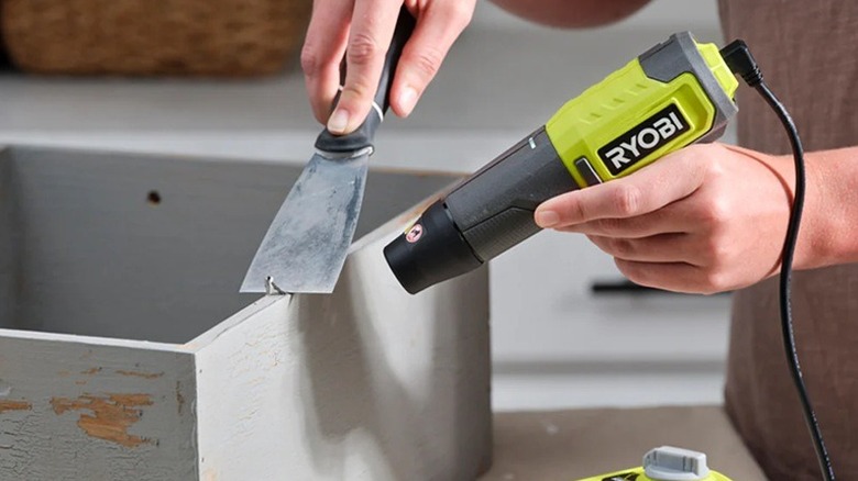 A person using a Ryobi heat pen and a putty knife to strip paint.