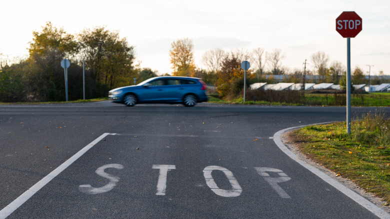 View of a road with a large white 'STOP' painted on the pavement, a stop sign on the right, and a blue car passing through the intersection.