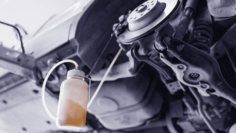 A brake bleeding process on a raised vehicle, showing a container partially filled with old brake fluid connected by a hose to the brake caliper.