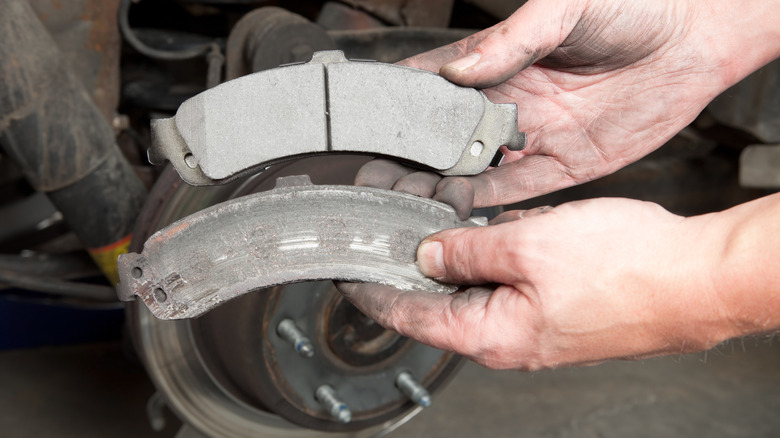 Mechanic holding a new brake pad above a severely worn brake pad in front of a car's exposed brake rotor during a brake service.