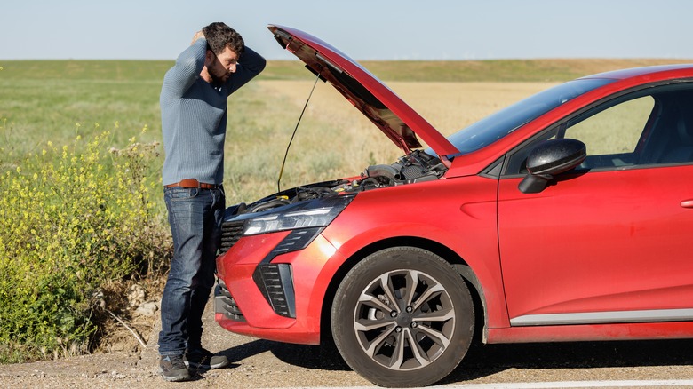 A man standing on the side of the road looking frustrated, holding his head while examining the engine of a red car with the hood open.