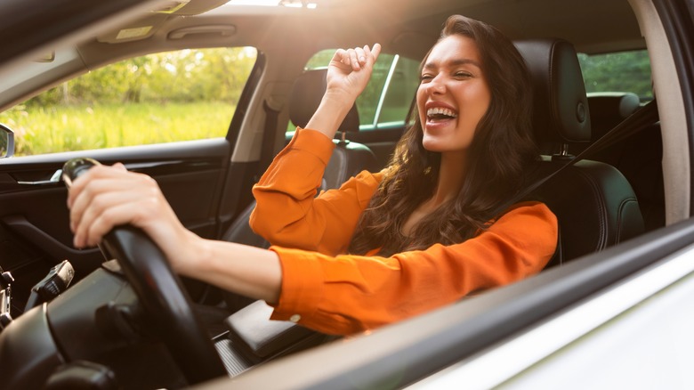 A smiling woman in an orange shirt driving a car, looking joyful with one hand on the steering wheel and the other raised in excitement.
