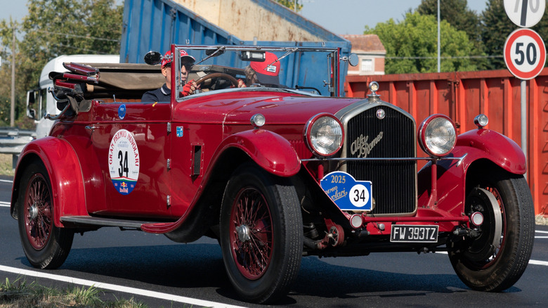 A red 1930s Alfa Romeo 6C Grand Touring roadster moving down a small road in the countryside.