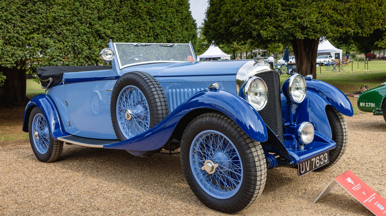 A baby blue Bentley 4.5 liter Dropehead Coupe coachbuilt touring car standing in front of manicured trees at a show.