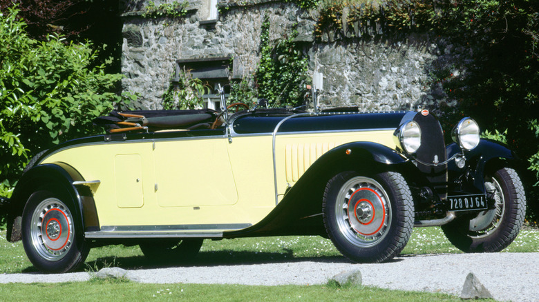 A cream colored 1934 Bugatti Type 46 standing against an old stone building on grass.