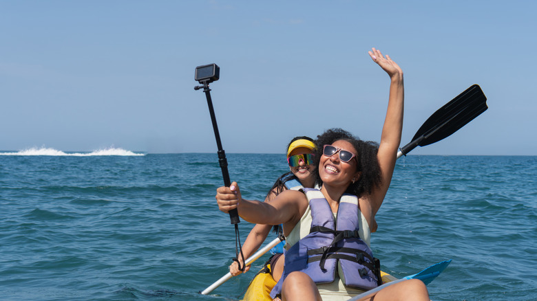 Two cheerful female tourists kayaking at sea and taking a selfie with action camera fixed on a stick